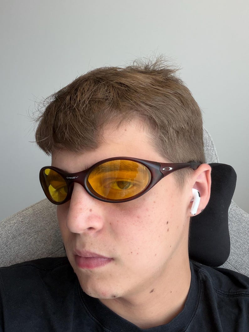 K&ouml;nnte beinhalten: Braune, ovale Sonnenbrille mit gelben Gl&auml;sern. Die Sonnenbrille hat einen dunkelbraunen Rahmen, und der Markenname "Oakley" ist oben auf dem Rahmen sichtbar. Die Gl&auml;ser reflektieren die Umgebung.