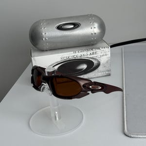 May include: Brown and black Oakley sunglasses with brown lenses, displayed on a clear stand. A silver case and a box with the text "SCIENCE AND ART" are in the background.