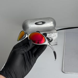Pu&ograve; includere: Occhiali da sole argentati con lenti a specchio rosse. Gli occhiali da sole hanno un design elegante e moderno. Una custodia argentata con il logo del marchio &egrave; dietro gli occhiali da sole. Le lenti riflettono una tonalit&agrave; arancione brillante. Gli occhiali da sole sono tenuti da una mano guantata.
