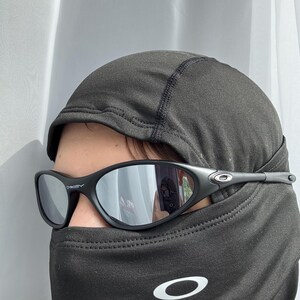 May include: Black Oakley sunglasses with mirrored lenses. The sunglasses are worn with a black balaclava. The Oakley logo is visible on the temples of the sunglasses and on the balaclava.