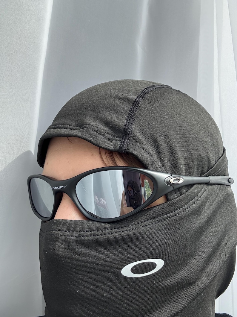 May include: Black Oakley sunglasses with mirrored lenses. The sunglasses are worn with a black balaclava. The Oakley logo is visible on the temples of the sunglasses and on the balaclava.