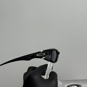 May include: Black sunglasses with a sleek, modern design. The sunglasses have a dark frame and lenses, with a white circular logo on the temples. The sunglasses are held up by a clear stand.
