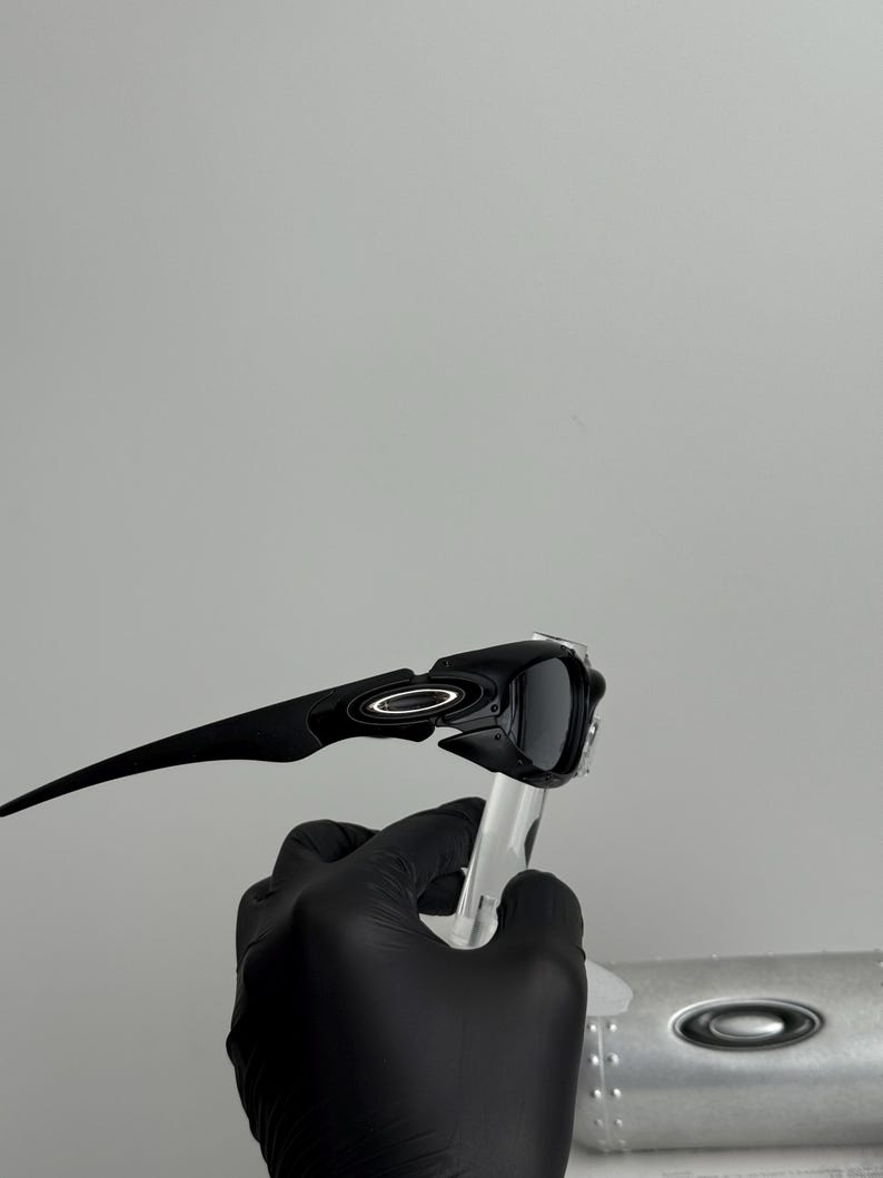 May include: Black sunglasses with a sleek, modern design. The sunglasses have a dark frame and lenses, with a white circular logo on the temples. The sunglasses are held up by a clear stand.