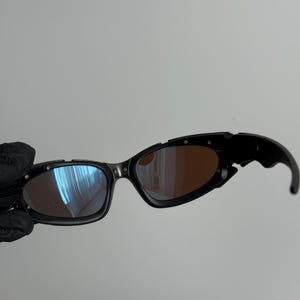 May include: Black sunglasses with a unique, angular design. The lenses have a mirrored finish, with a blue hue on one side and a brown tint on the other. The frames are black with silver accents.