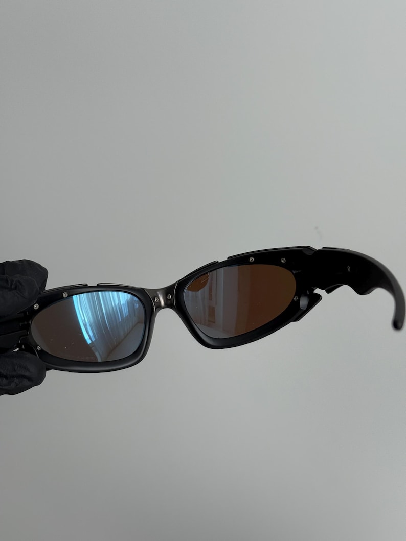 May include: Black sunglasses with a unique, angular design. The lenses have a mirrored finish, with a blue hue on one side and a brown tint on the other. The frames are black with silver accents.