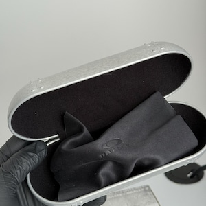 K&ouml;nnte beinhalten: Ge&ouml;ffnete silberfarbene Brillenetui mit schwarzem Innenraum und schwarzem Mikrofasertuch. Das Etui hat ein genietetes Design und das Oakley-Logo ist auf dem Tuch sichtbar.
