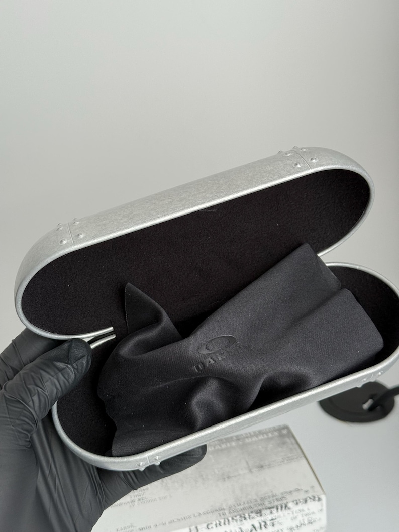 K&ouml;nnte beinhalten: Ge&ouml;ffnete silberfarbene Brillenetui mit schwarzem Innenraum und schwarzem Mikrofasertuch. Das Etui hat ein genietetes Design und das Oakley-Logo ist auf dem Tuch sichtbar.