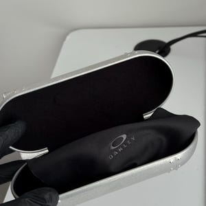 May include: Open silver-colored Oakley glasses case with a black interior and a black microfiber cloth. The Oakley logo is visible on the cloth. The case is open, revealing the interior.