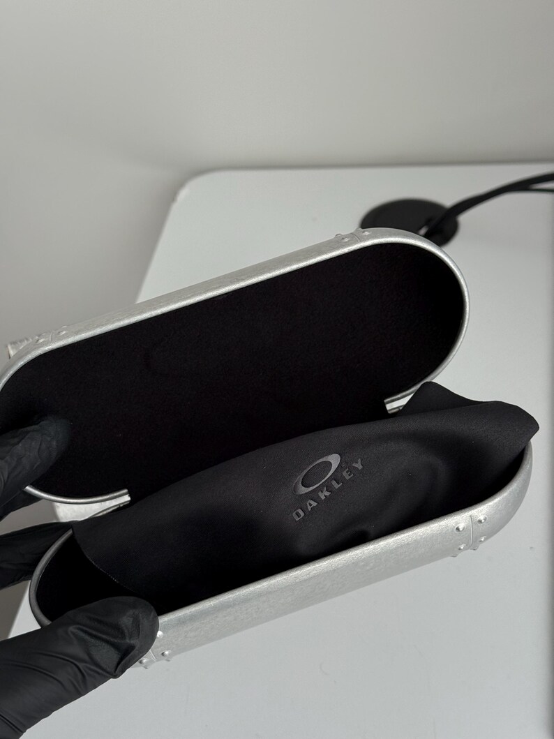 May include: Open silver-colored Oakley glasses case with a black interior and a black microfiber cloth. The Oakley logo is visible on the cloth. The case is open, revealing the interior.
