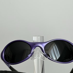 May include: Purple sunglasses with dark lenses. The sunglasses have a rounded frame and a clear nose bridge. The word "SOLS" is printed on the top of the frame. The sunglasses are held by a clear display stand.