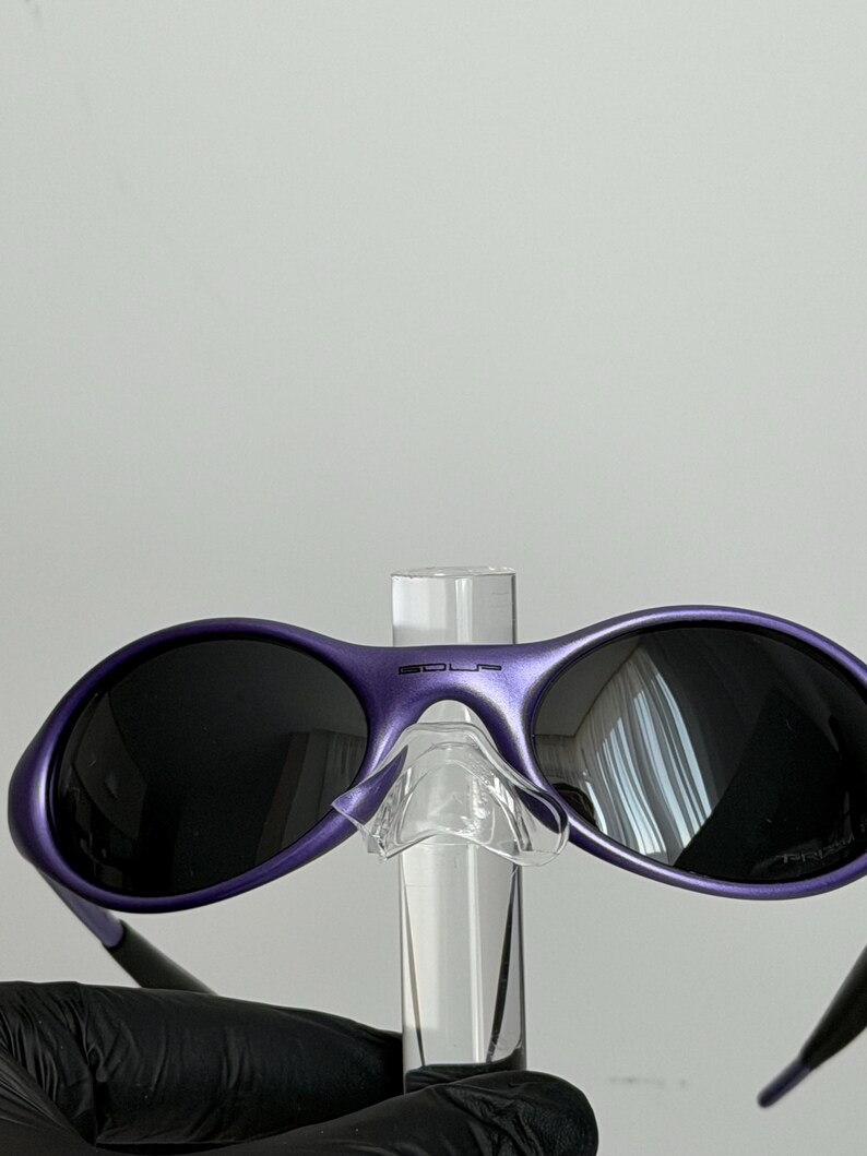 May include: Purple sunglasses with dark lenses. The sunglasses have a rounded frame and a clear nose bridge. The word "SOLS" is printed on the top of the frame. The sunglasses are held by a clear display stand.