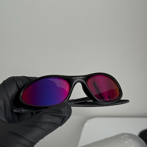 May include: Black sunglasses with a curved frame and reflective lenses that transition from purple to red. The brand name "Oakley" is visible on the frame. The sunglasses are held by a gloved hand.