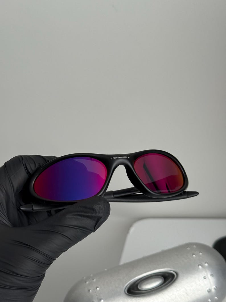 May include: Black sunglasses with a curved frame and reflective lenses that transition from purple to red. The brand name "Oakley" is visible on the frame. The sunglasses are held by a gloved hand.