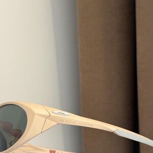 May include: Beige-colored sunglasses with dark lenses. The sunglasses have a sleek, modern design with a curved frame and white accents on the arms. The text "RN 98548-CA35460" is printed on the arm.