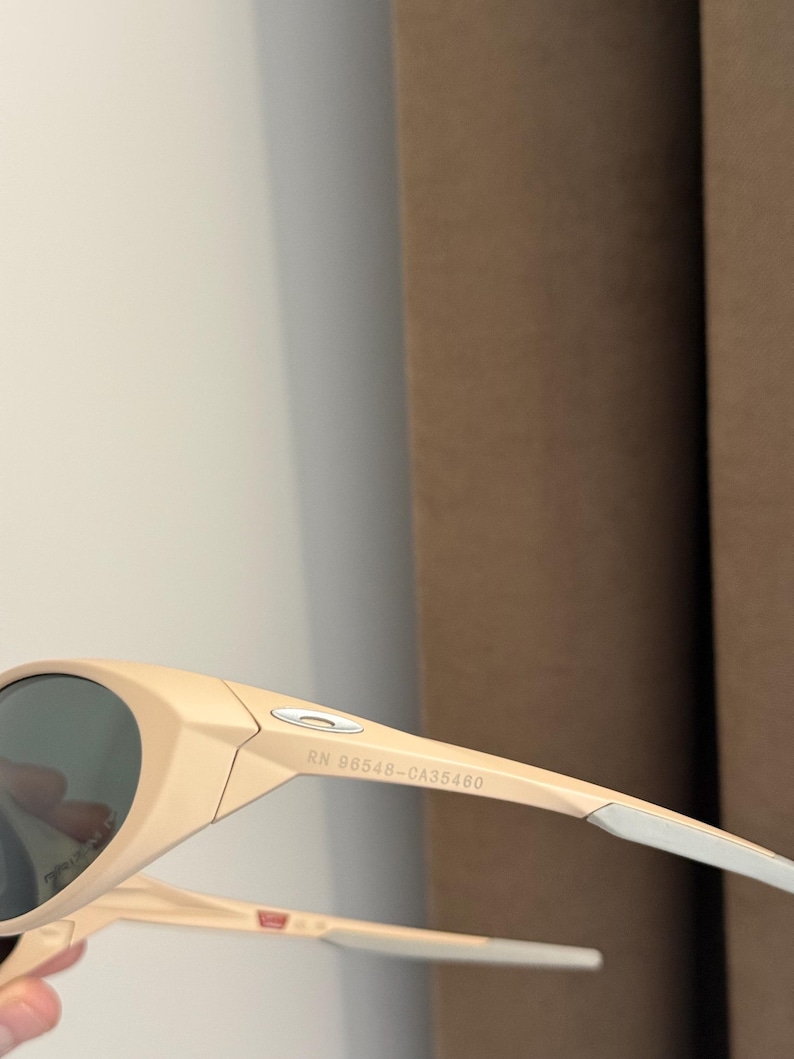 May include: Beige-colored sunglasses with dark lenses. The sunglasses have a sleek, modern design with a curved frame and white accents on the arms. The text "RN 98548-CA35460" is printed on the arm.