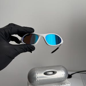 May include: White-framed sunglasses with blue mirrored lenses. The sunglasses are held by a gloved hand. A silver case and a box with the text "SCIENCE AND ART" are in the background.