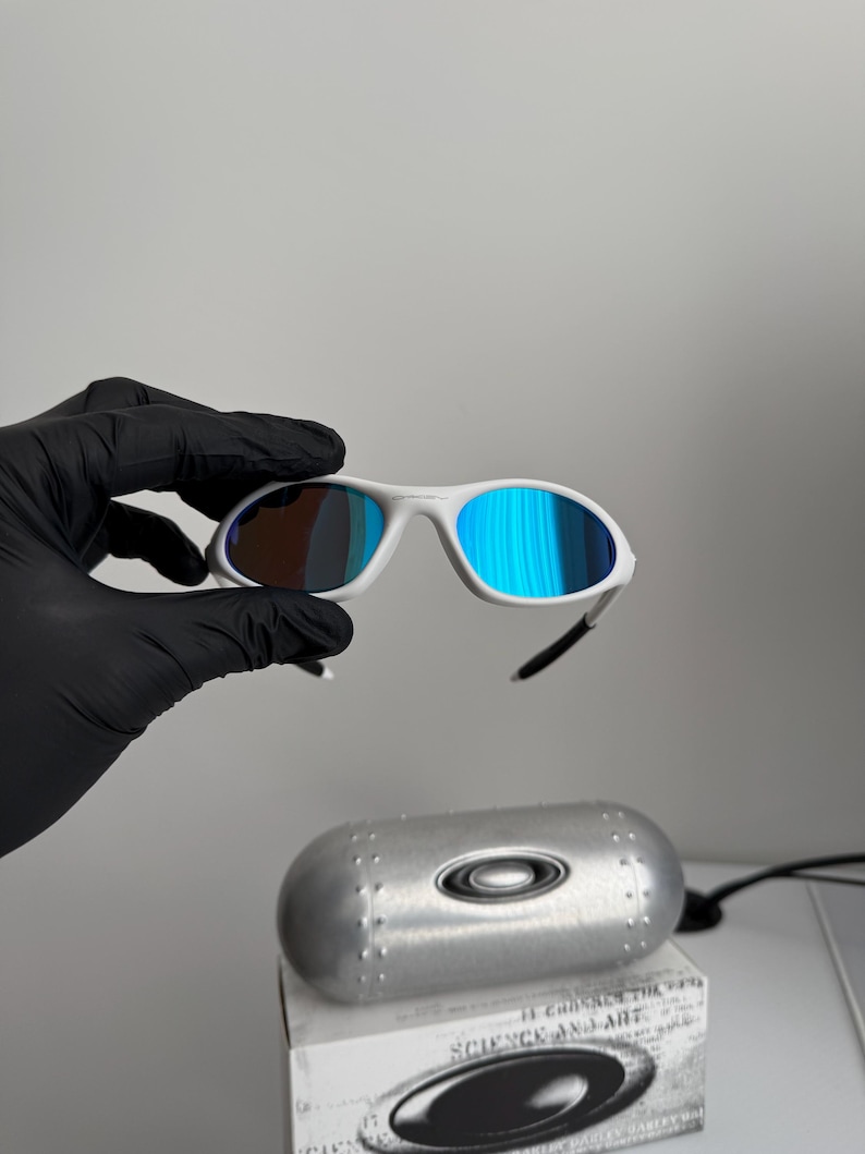 May include: White-framed sunglasses with blue mirrored lenses. The sunglasses are held by a gloved hand. A silver case and a box with the text "SCIENCE AND ART" are in the background.