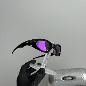 May include: Black sunglasses with purple mirrored lenses are displayed on a clear stand. The sunglasses have a sleek, sporty design. A silver case and a box with the text "SCIENCE AND ART" are in the background.