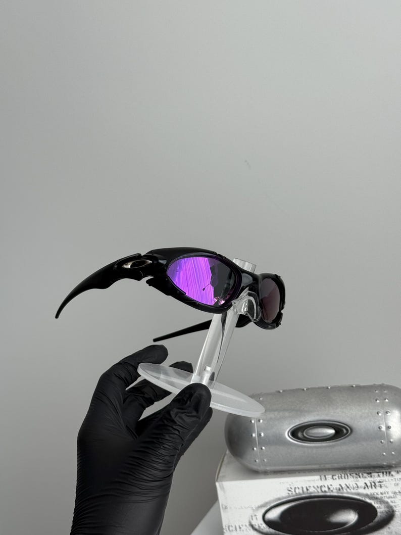 May include: Black sunglasses with purple mirrored lenses are displayed on a clear stand. The sunglasses have a sleek, sporty design. A silver case and a box with the text "SCIENCE AND ART" are in the background.