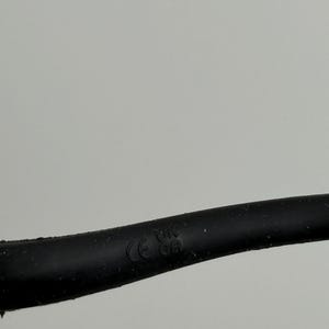 Puede incluir: Primer plano de un cable el&eacute;ctrico negro con la inscripci&oacute;n "CE UK CA". El cable est&aacute; ligeramente curvado sobre un fondo gris claro liso.
