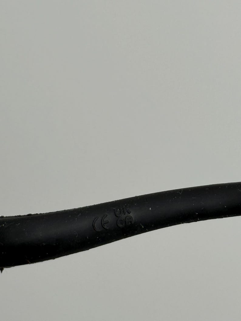 Puede incluir: Primer plano de un cable el&eacute;ctrico negro con la inscripci&oacute;n "CE UK CA". El cable est&aacute; ligeramente curvado sobre un fondo gris claro liso.