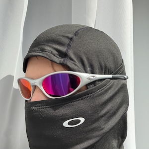 May include: Silver sunglasses with red and purple mirrored lenses. The sunglasses are worn with a black balaclava featuring a white Oakley logo. The balaclava covers the lower face and neck.