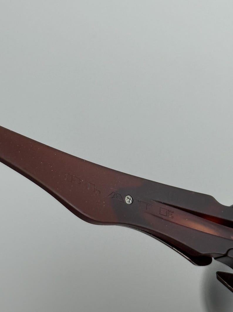 May include: Close-up of a brown and black patterned eyeglass arm. The arm has text and a small silver screw. The glasses are set against a light gray background.