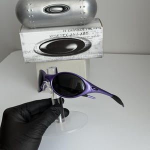May include: Purple sunglasses with black lenses are displayed on a clear stand. A silver case and a white box with the text "SCIENCE AND ART" are in the background. The sunglasses have a sleek, modern design.