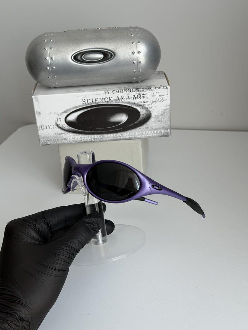 May include: Purple sunglasses with black lenses are displayed on a clear stand. A silver case and a white box with the text "SCIENCE AND ART" are in the background. The sunglasses have a sleek, modern design.
