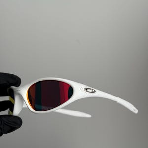 May include: White sunglasses with a curved frame and red-tinted lenses. The sunglasses have a sleek, modern design with the brand logo on the temples. The lenses reflect a gradient of colours.