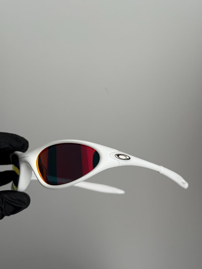 May include: White sunglasses with a curved frame and red-tinted lenses. The sunglasses have a sleek, modern design with the brand logo on the temples. The lenses reflect a gradient of colours.
