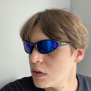 Puede incluir: Gafas de sol negras con lentes azules reflectantes. Las gafas tienen una montura curvada y un diseño deportivo. Las lentes reflejan un color azul brillante. Un accesorio popular para actividades al aire libre.