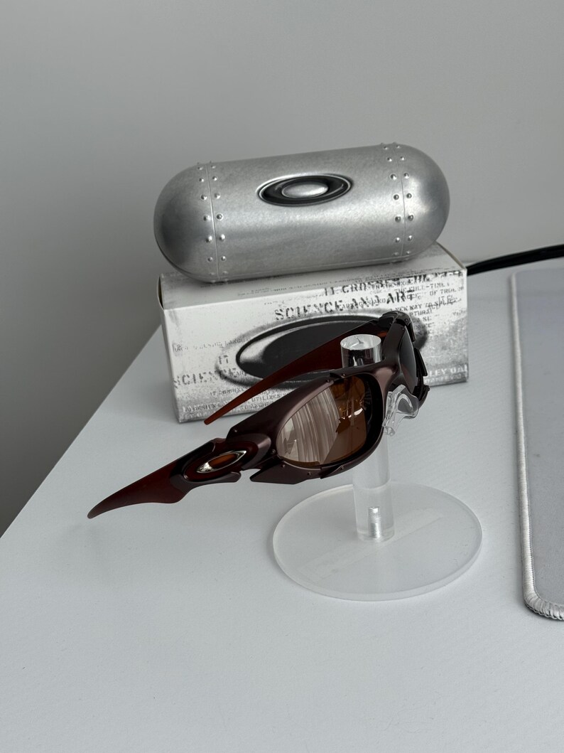 May include: Brown and black sunglasses with a unique, angular design, displayed on a clear stand. A silver case and a box with text are in the background. The sunglasses have brown lenses and a distinctive frame.