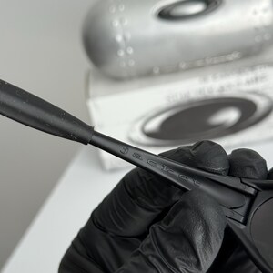 May include: Black sunglasses with the word "jacket" printed on the arm. The sunglasses are held by a gloved hand. A silver case and a white box are in the background.