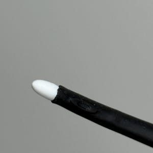 Puede incluir: Primer plano de un aplicador negro con una punta blanca y redondeada. El aplicador est&aacute; en &aacute;ngulo diagonal sobre un fondo gris. La punta parece estar hecha de un material suave y absorbente.