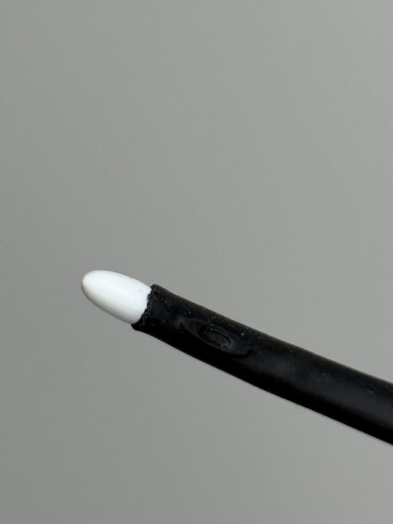 Puede incluir: Primer plano de un aplicador negro con una punta blanca y redondeada. El aplicador est&aacute; en &aacute;ngulo diagonal sobre un fondo gris. La punta parece estar hecha de un material suave y absorbente.