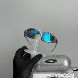 May include: Silver-framed sunglasses with blue lenses are displayed on a clear stand. The sunglasses are set against a gray background, with a silver case and a box with the text "SCIENCE AND ART" visible.