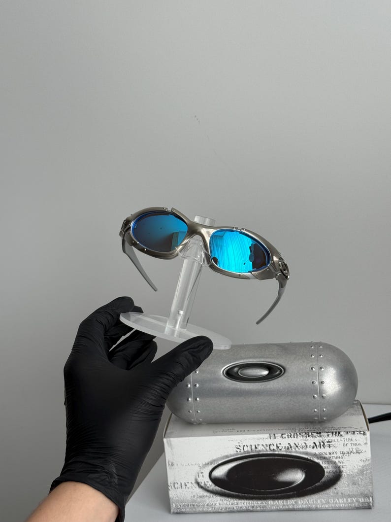 May include: Silver-framed sunglasses with blue lenses are displayed on a clear stand. The sunglasses are set against a gray background, with a silver case and a box with the text "SCIENCE AND ART" visible.