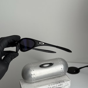 May include: Black sunglasses with a curved frame and dark lenses. The sunglasses are displayed with a silver case and a box with the text "SCIENCE AND ART". The Oakley logo is visible on the sunglasses.