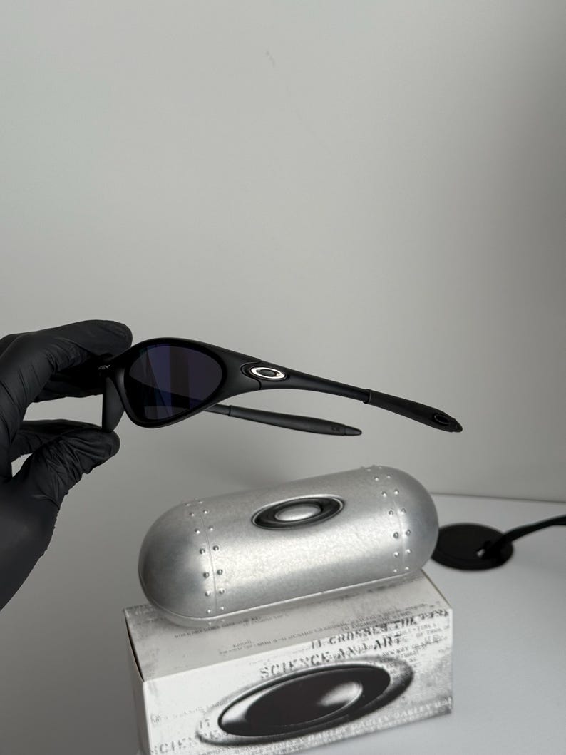 May include: Black sunglasses with a curved frame and dark lenses. The sunglasses are displayed with a silver case and a box with the text "SCIENCE AND ART". The Oakley logo is visible on the sunglasses.