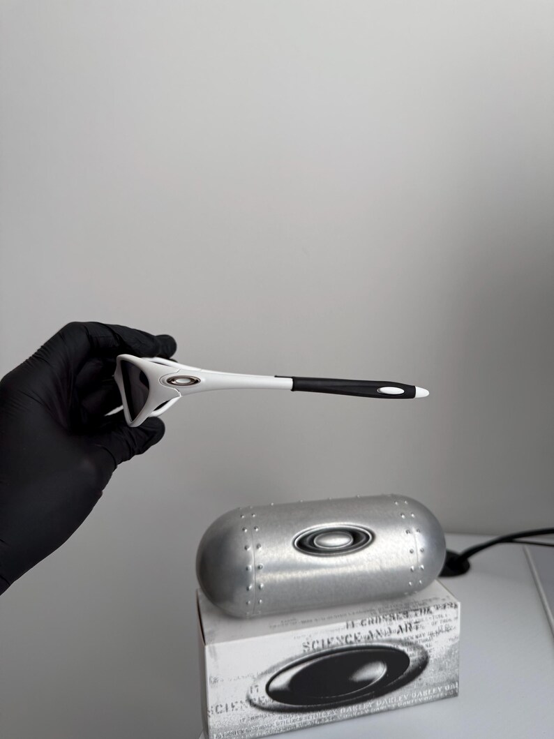 May include: White and black sports sunglasses with a silver case and a box. The sunglasses have a unique design with a white frame and black accents. The case is metallic silver with a matching logo.