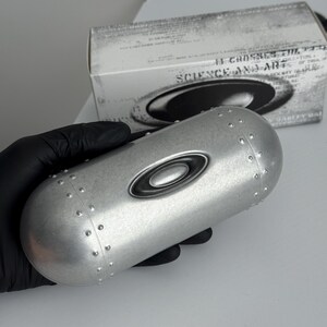May include: A silver-colored eyewear case with the Oakley logo, held by a gloved hand. The case has a cylindrical shape with rivets and a black oval logo. A box with the text "SCIENCE AND ART" is in the background.