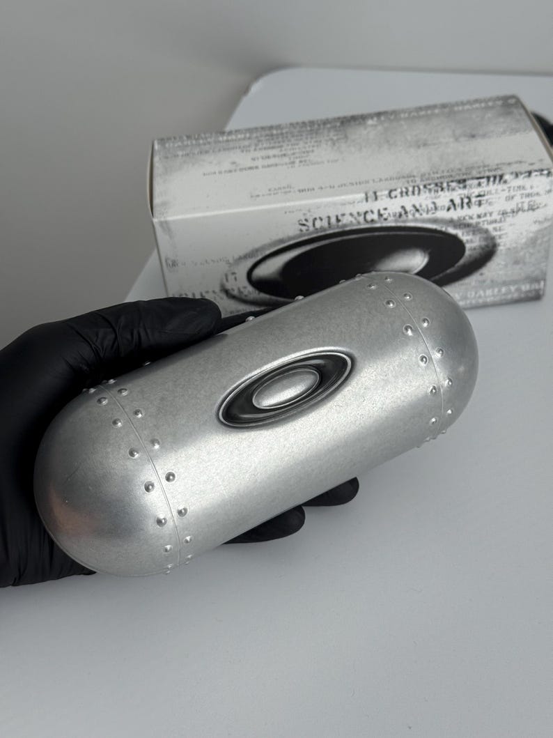 May include: A silver-colored eyewear case with the Oakley logo, held by a gloved hand. The case has a cylindrical shape with rivets and a black oval logo. A box with the text "SCIENCE AND ART" is in the background.