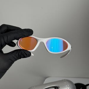 May include: White-framed sunglasses with orange and blue-tinted lenses. The sunglasses have a sleek, modern design. The lenses reflect a gradient of colors. The sunglasses are shown with a silver case.