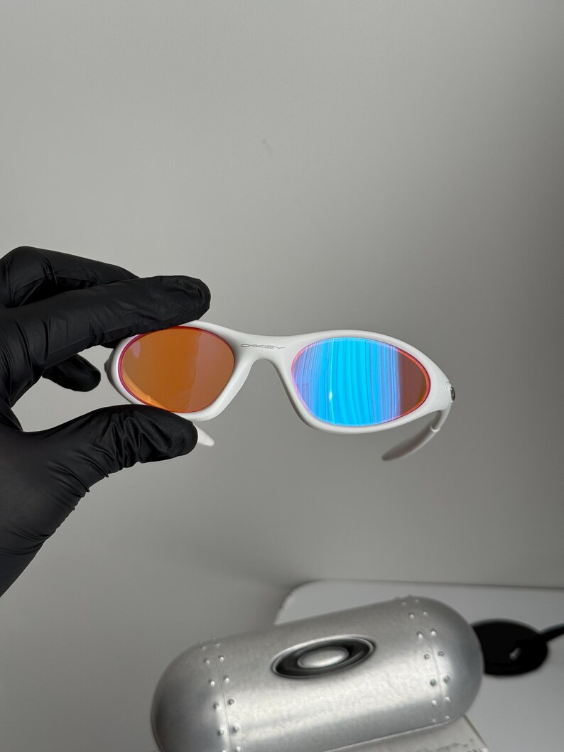 May include: White-framed sunglasses with orange and blue-tinted lenses. The sunglasses have a sleek, modern design. The lenses reflect a gradient of colors. The sunglasses are shown with a silver case.