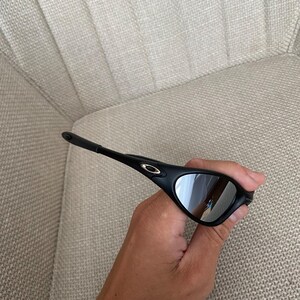Puede incluir: Gafas de sol negras con lentes espejadas plateadas. Las gafas tienen un dise&ntilde;o elegante y moderno con una montura curvada y un peque&ntilde;o logotipo dorado en la patilla. Las lentes reflejan el entorno.