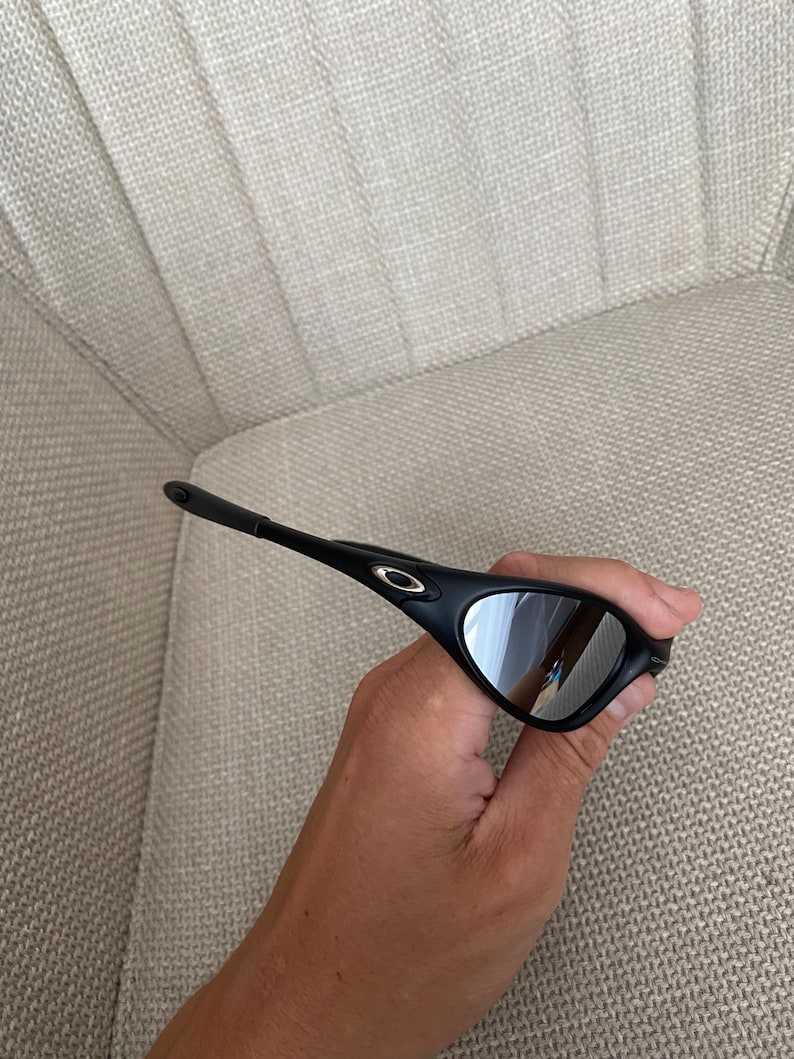 Puede incluir: Gafas de sol negras con lentes espejadas plateadas. Las gafas tienen un dise&ntilde;o elegante y moderno con una montura curvada y un peque&ntilde;o logotipo dorado en la patilla. Las lentes reflejan el entorno.