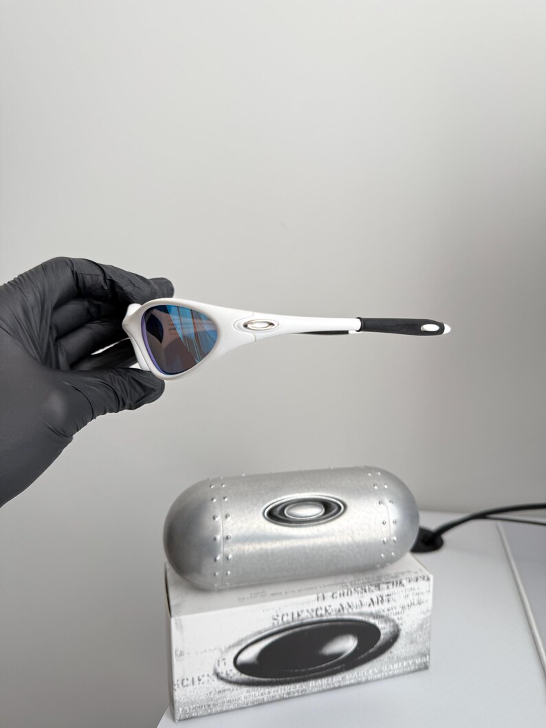 May include: White sunglasses with blue lenses are held by a gloved hand. The sunglasses have a sleek design with black accents. A silver case and a box with the text "SCIENCE AND ART" are also visible.