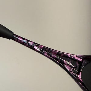May include: Close-up of a black and purple patterned sunglasses arm. The arm has a speckled design with the brand name etched in silver. The end of the arm is black.
