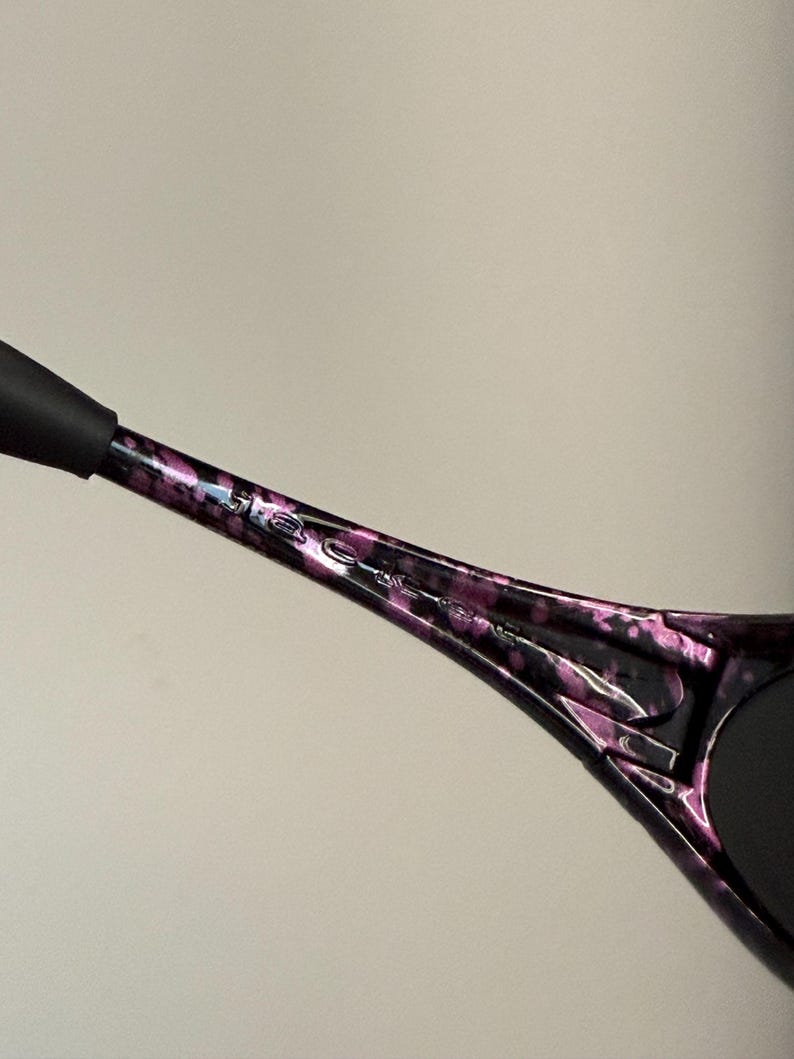 May include: Close-up of a black and purple patterned sunglasses arm. The arm has a speckled design with the brand name etched in silver. The end of the arm is black.